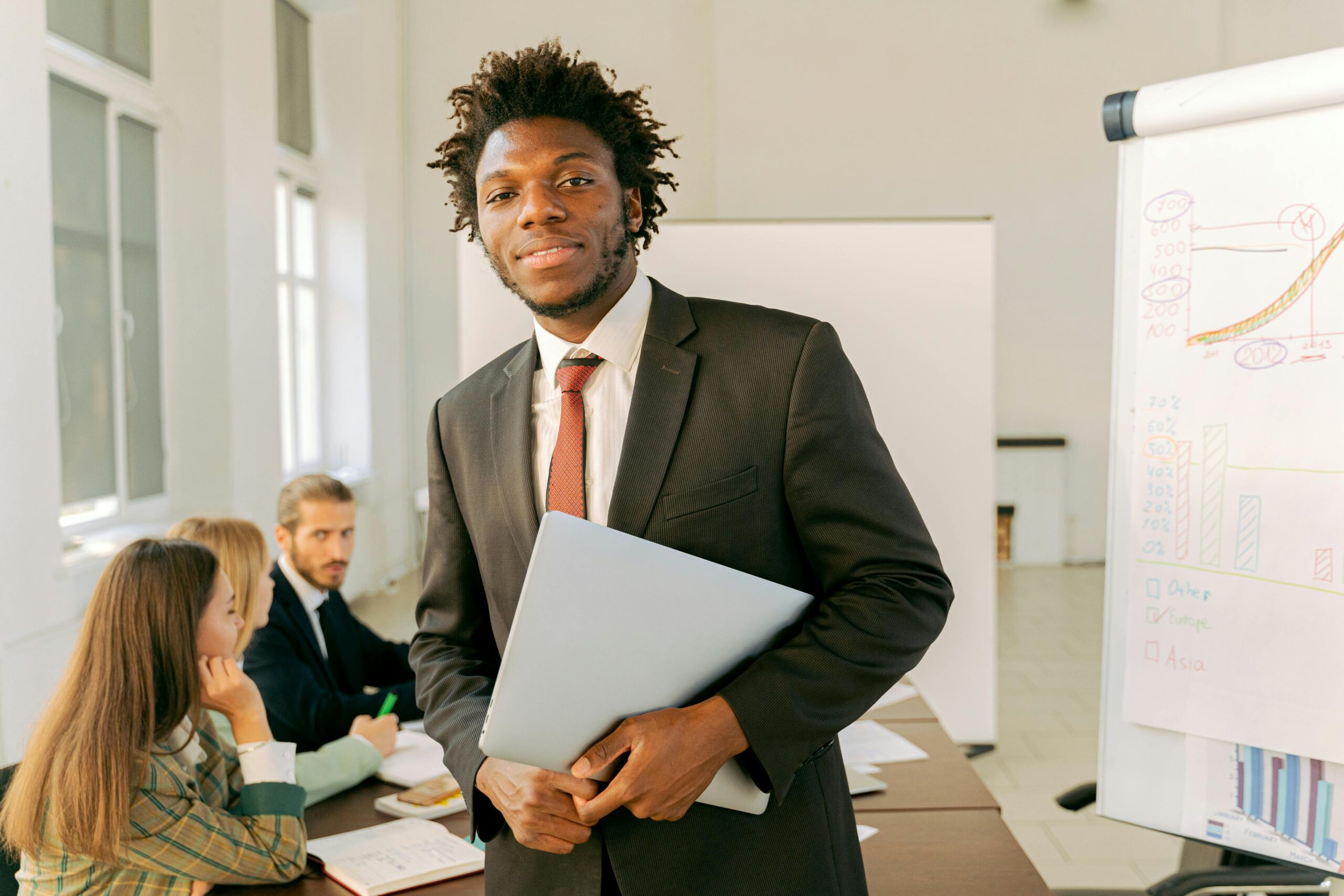 A confident businessman in a black suit holds a laptop during a meeting in an office setting.
